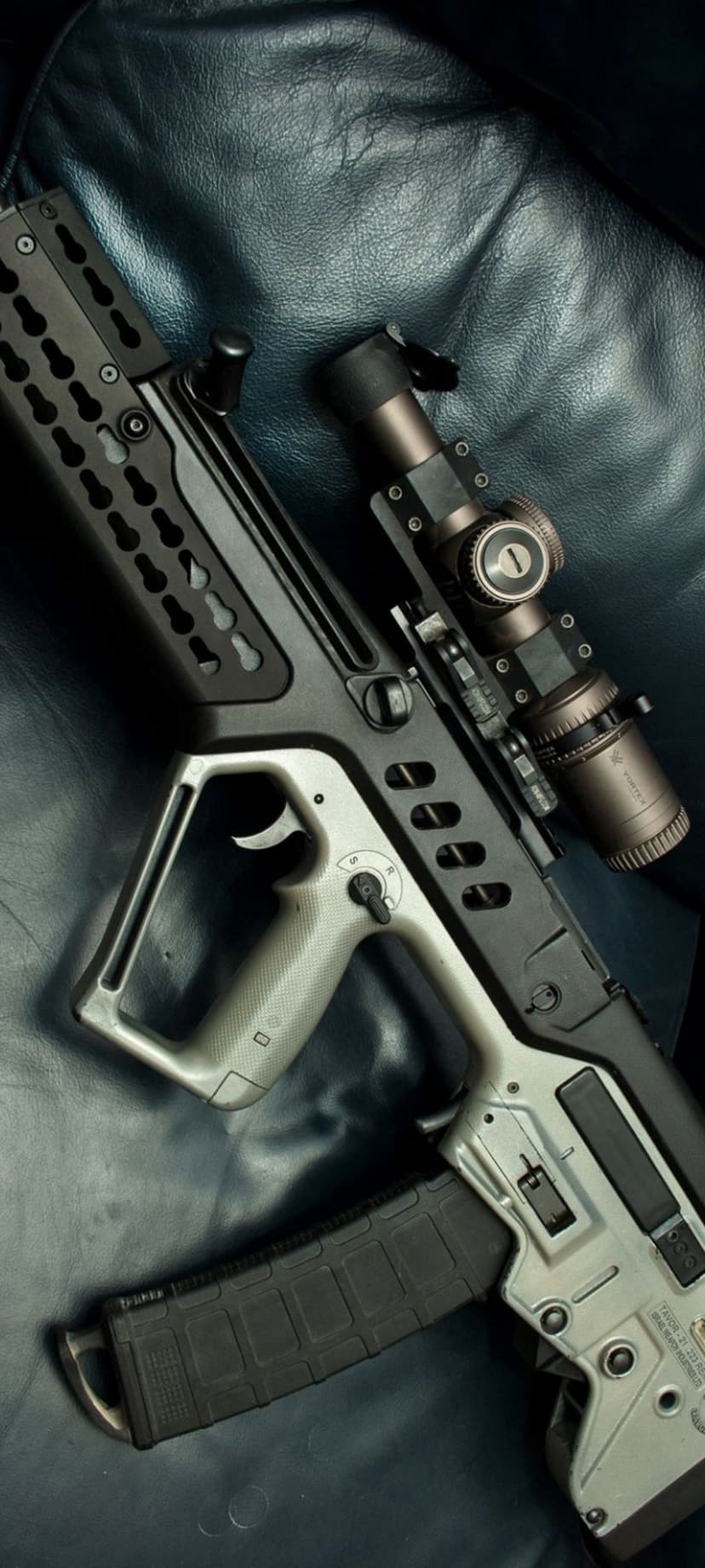 Modern bullpup rifle with a top-mounted scope and magazine laying on a dark textured leather surface.