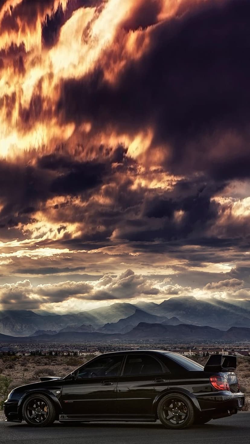 Black Subaru Impreza sedan parked on a road with a dramatic orange sunset and mountain peaks in the background.