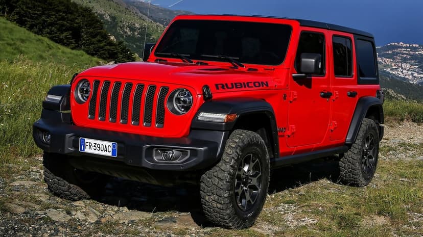 Red Jeep Wrangler Rubicon parked on a rocky green mountain ridge under a bright blue sky with a distant city.