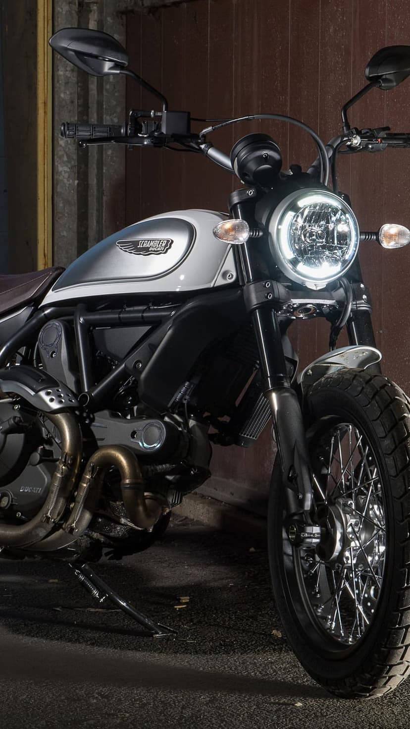 Ducati Scrambler motorcycle featuring a glowing circular LED headlight, silver fuel tank, and spoked front wheel.