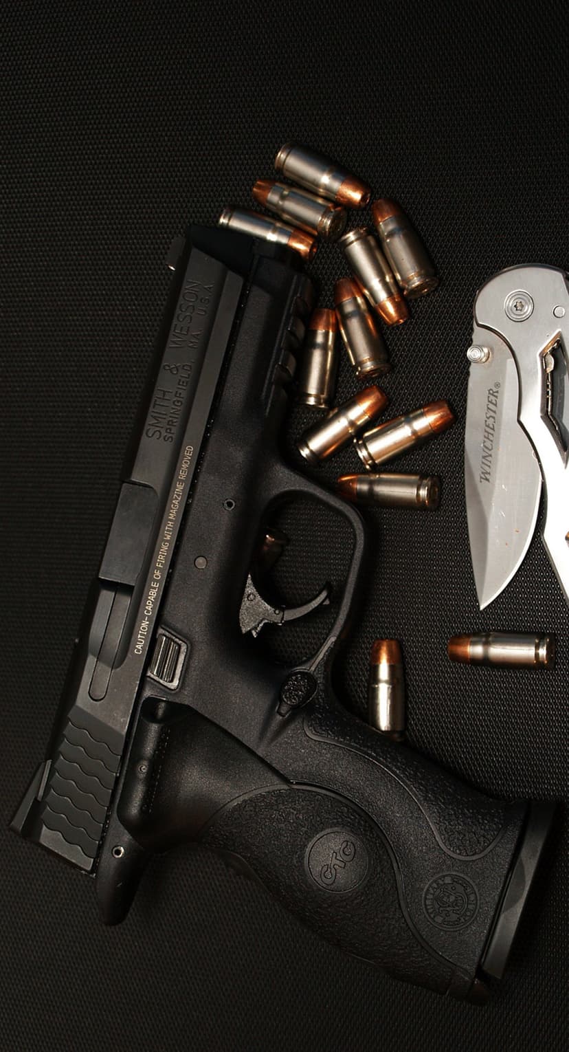 Smith & Wesson handgun, scattered brass ammunition, and a silver folding knife rest on a dark textured surface.