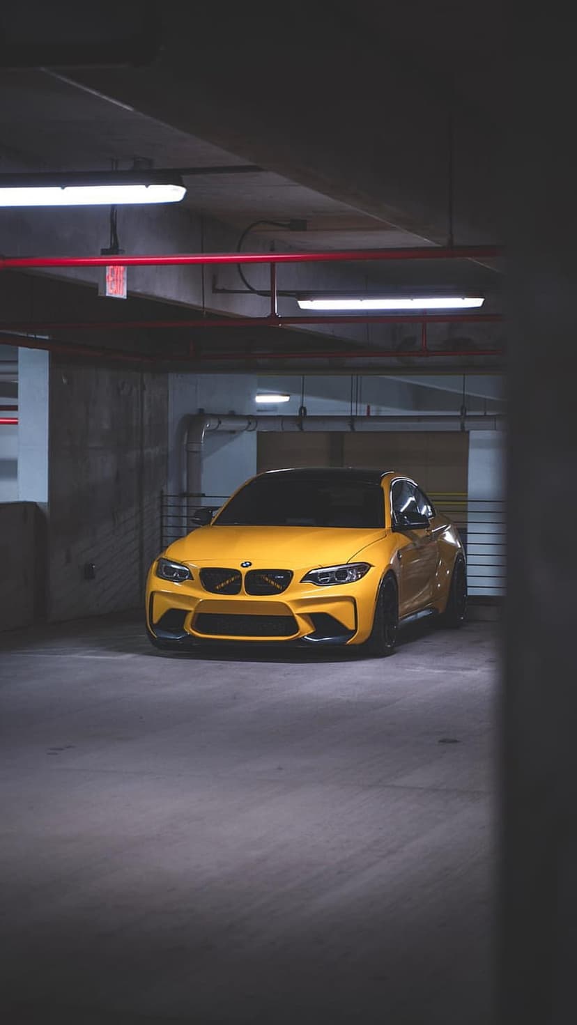 Yellow BMW M2 coupe parked in a dark concrete garage with exposed pipes and dim fluorescent overhead lighting.