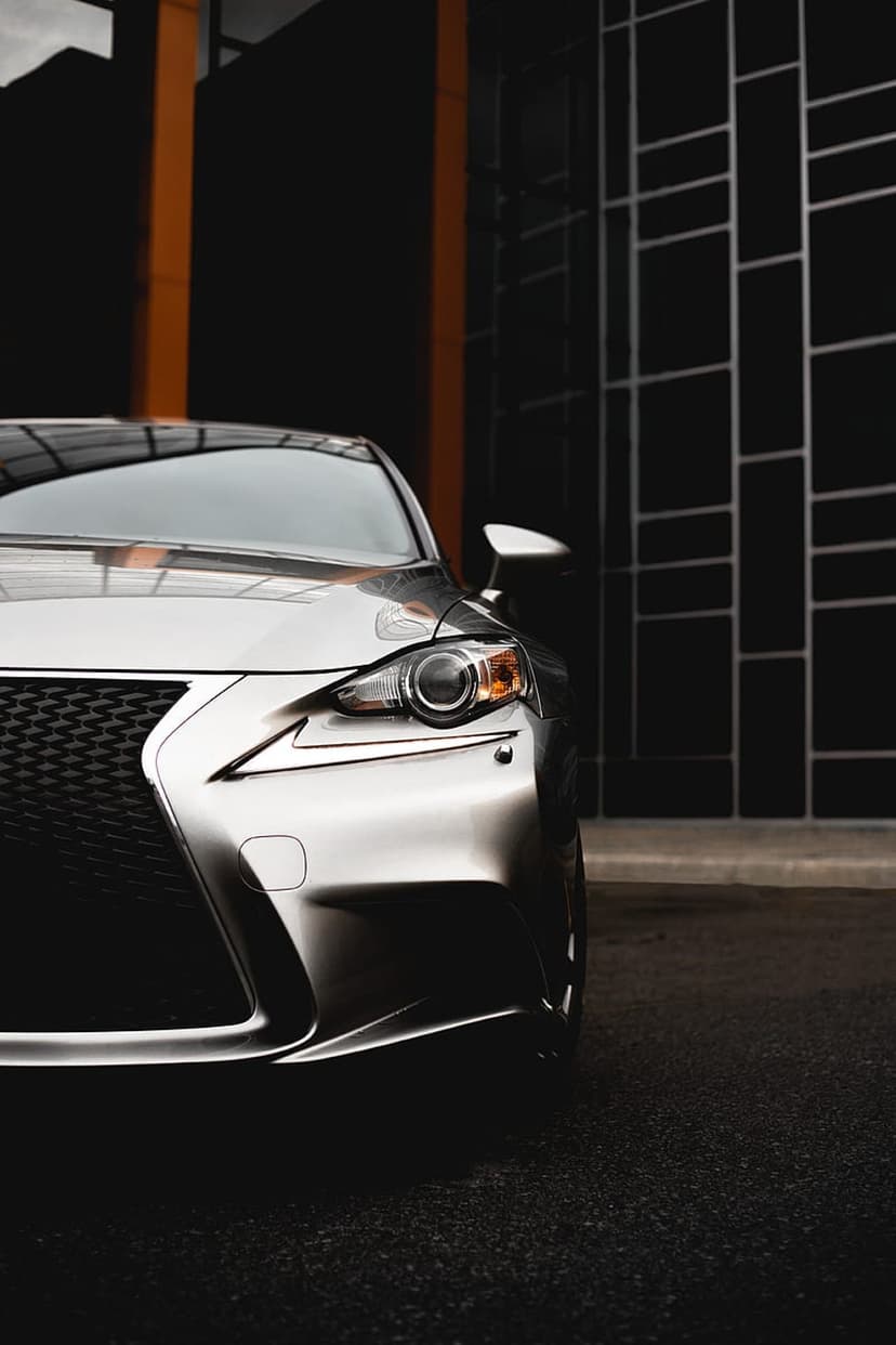 Silver Lexus IS front grille and headlights against a black tiled wall with orange accents in a low-angle shot.