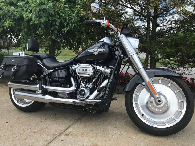 Black Harley-Davidson Fat Boy 114 motorcycle with chrome engine and solid disc wheels parked by green trees.