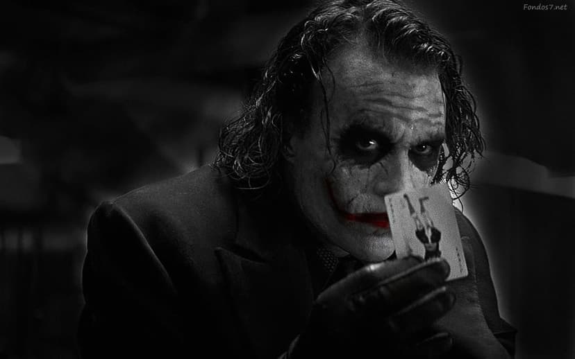 Heath Ledger as Joker holds a playing card featuring a hanging man silhouette against a dark blurred background.