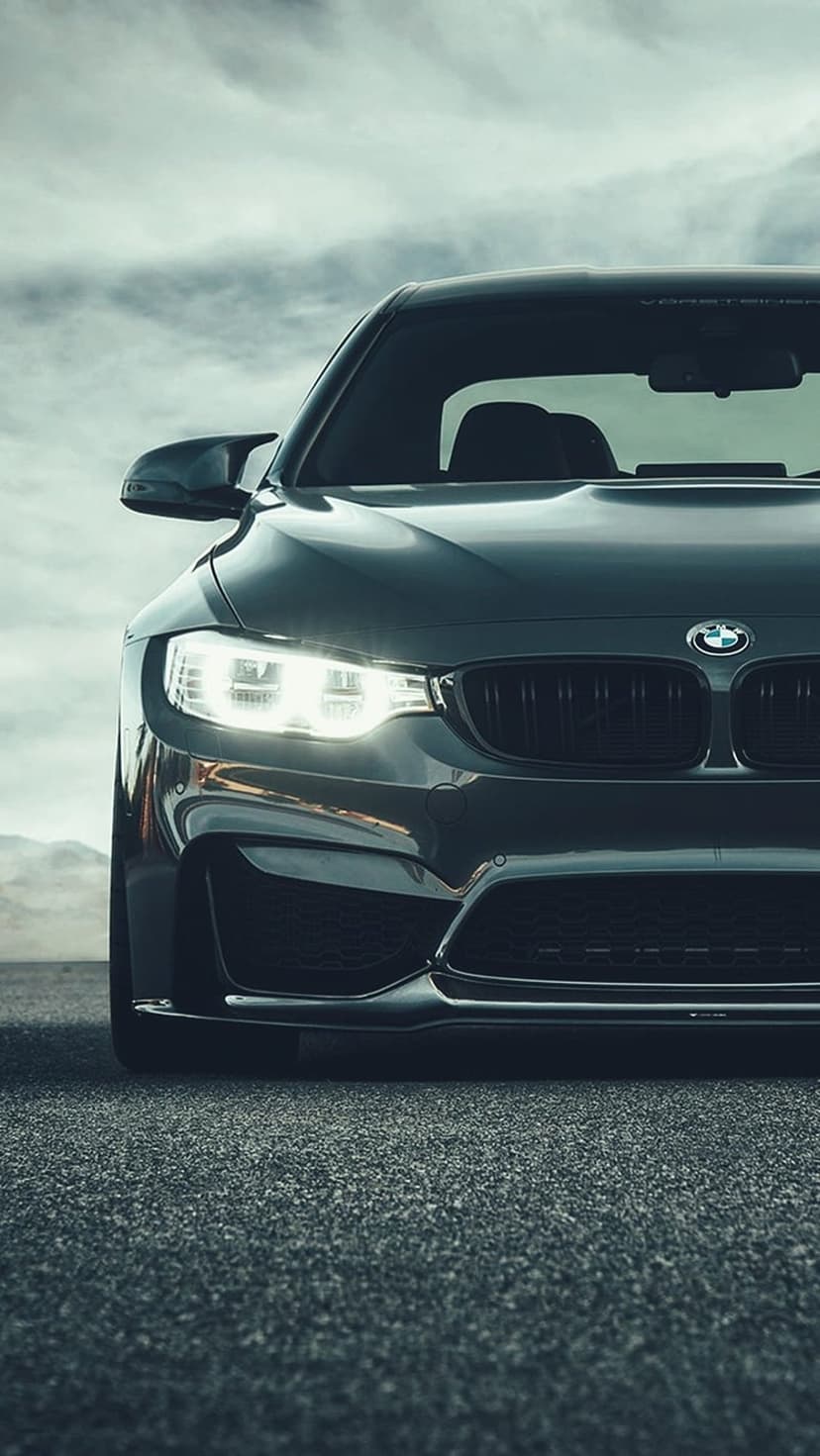 Dark gray BMW M4 with glowing LED headlights parked on asphalt against a cloudy sky and mountain backdrop.