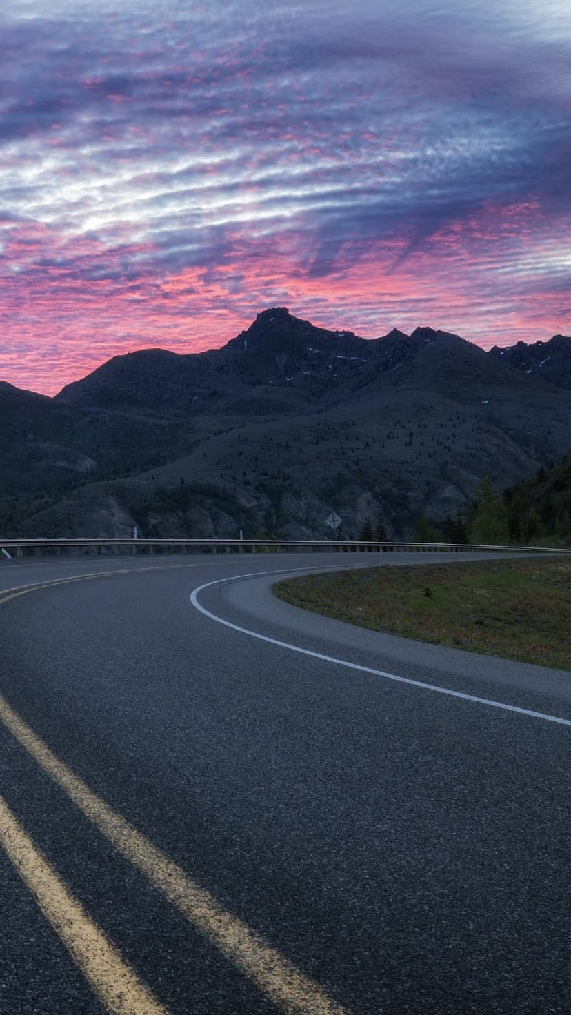 Winding asphalt road curves through dark mountain silhouettes under a glowing pink and purple twilight sky.