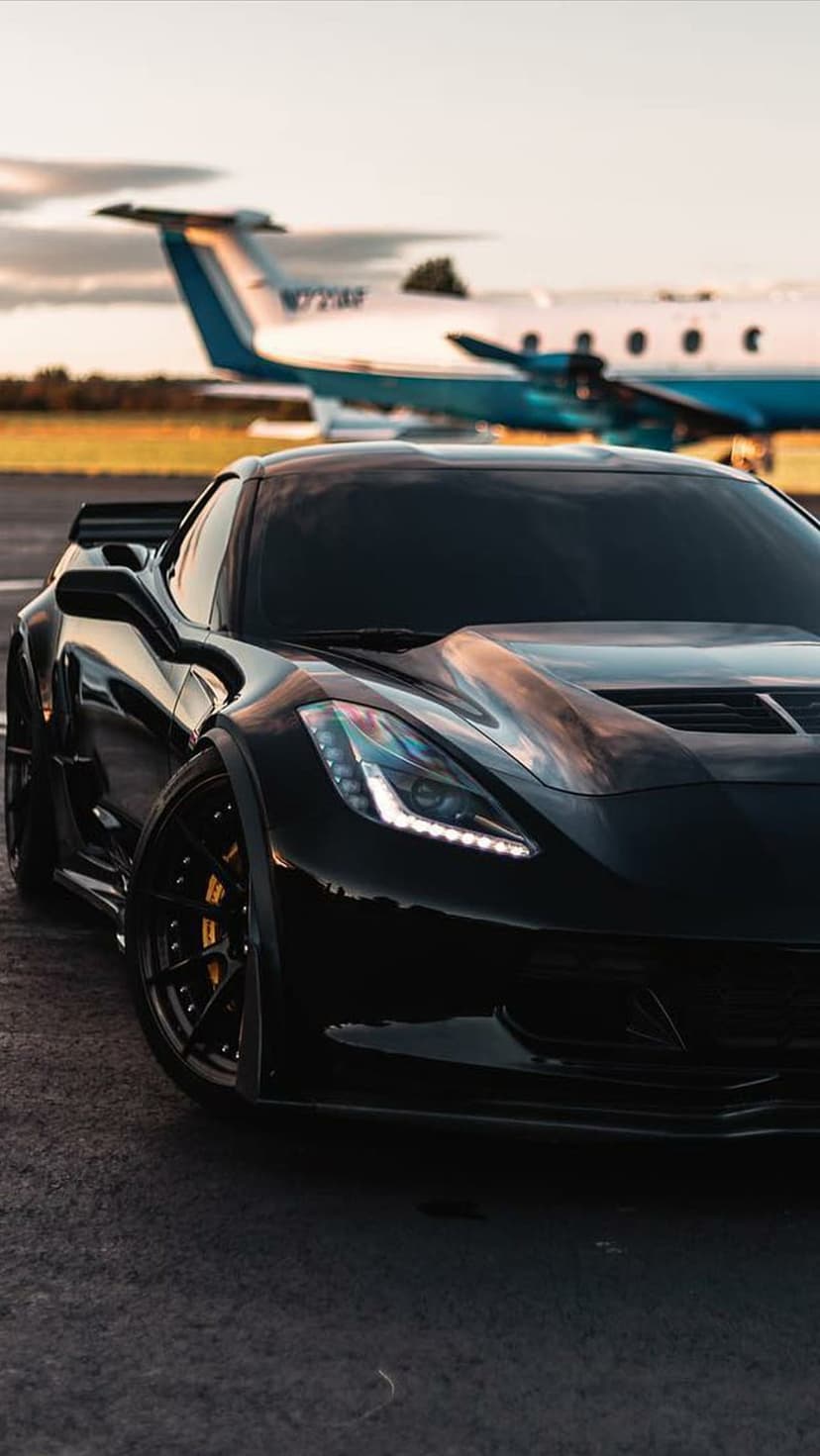 Black Corvette parked next to a private jet on a tarmac during golden hour with a warm sunset sky in the background.