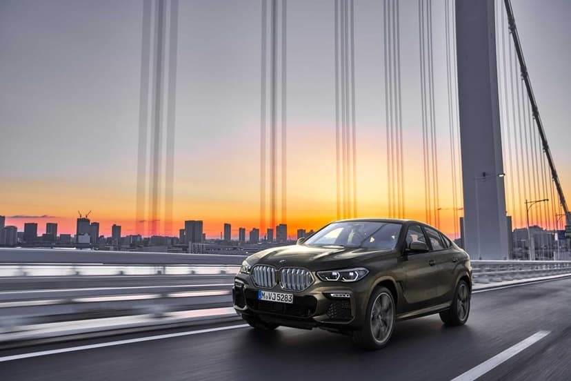 Bronze BMW X6 driving on a suspension bridge with a sunset sky and city skyline in the background.