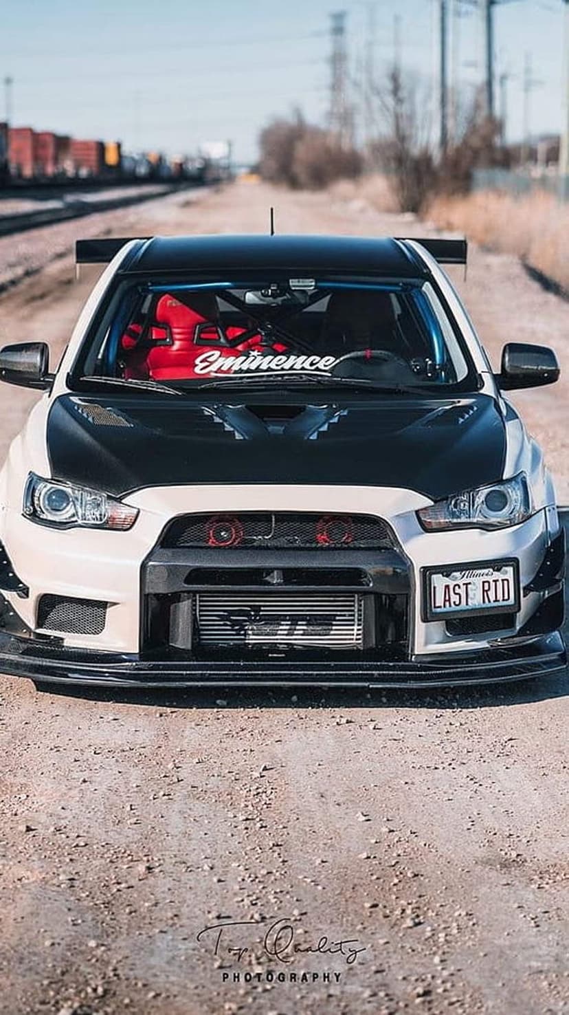 White Mitsubishi Evo X with a rear wing and intercooler parked on a dirt path with a train in the distance.