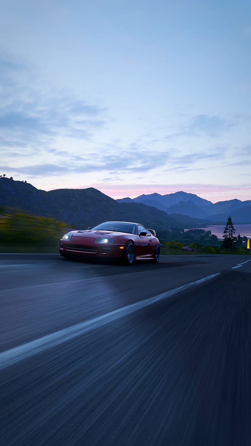Red Toyota Supra driving fast on a mountain highway with a lake and pink twilight sky in the background.