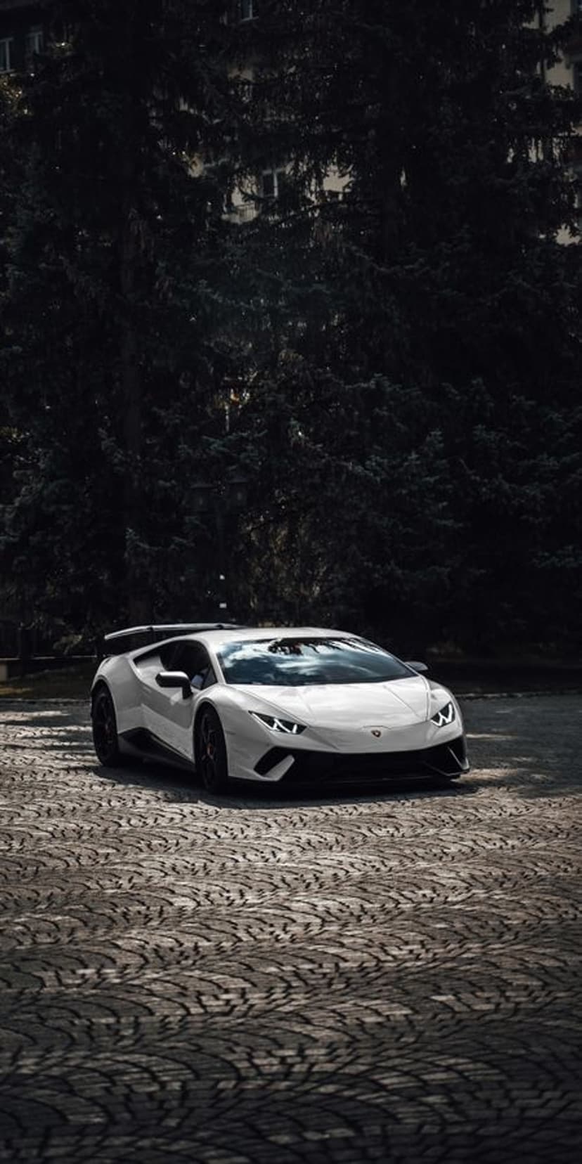 White Lamborghini Huracan Evo parked on patterned cobblestones with dark trees and soft outdoor lighting.