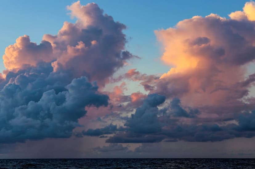 Billowing orange and pink clouds tower over a dark, calm sea during a dramatic sunset at the horizon.