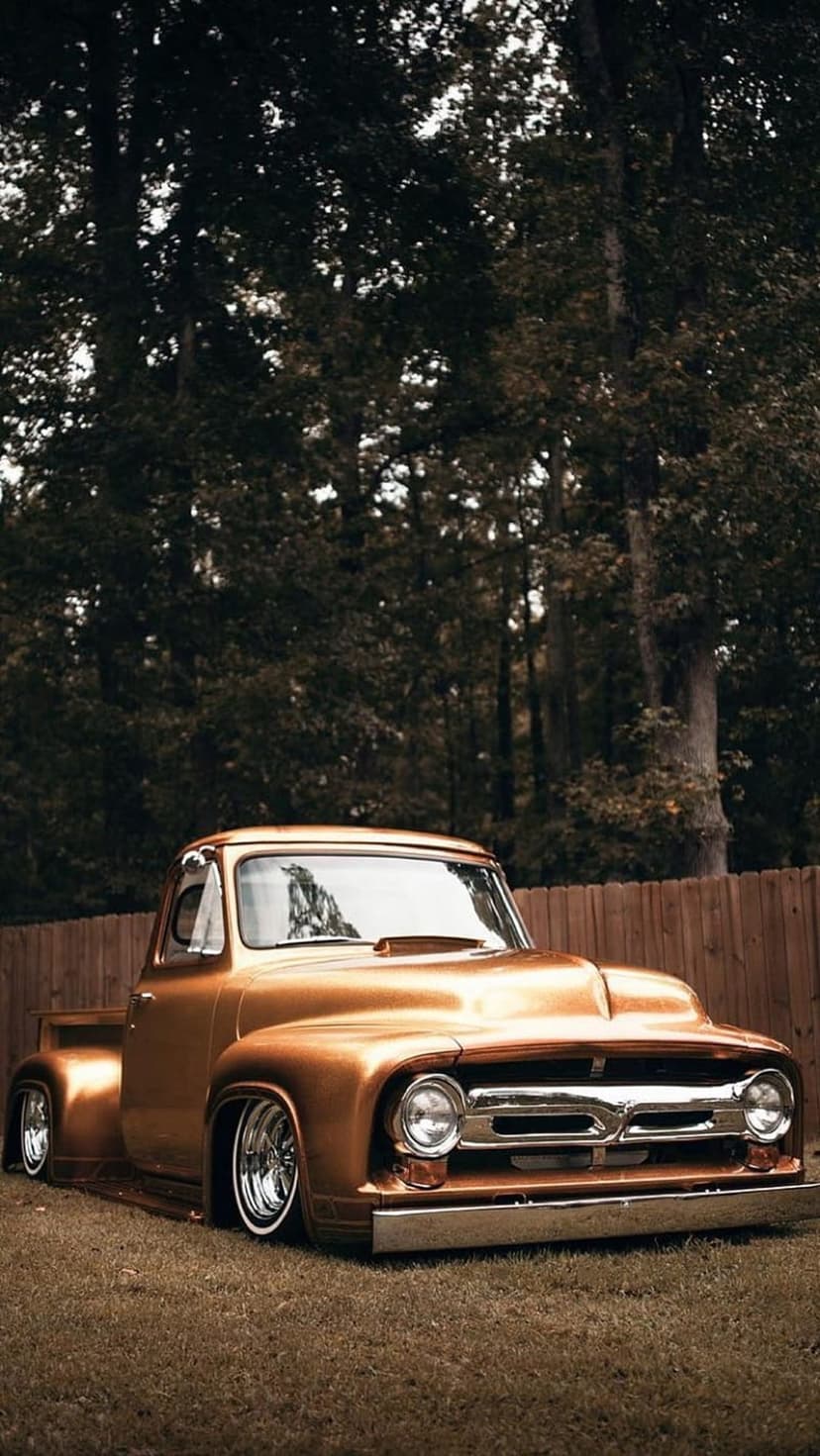 Copper vintage Ford truck with chrome wheels parked on grass near a wooden fence in warm afternoon sunlight.