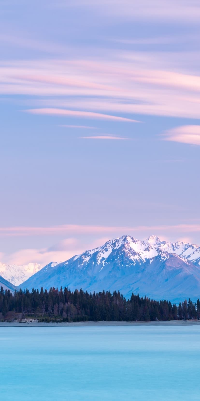 Snow-capped peaks tower over a blue lake and dark pine forest under a soft pink and purple sunset sky.