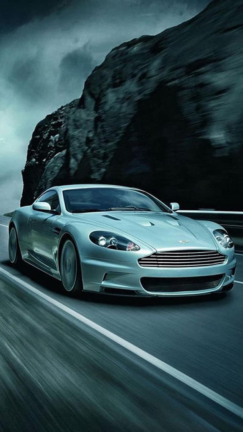 Silver Aston Martin DB9 speeds past a rocky cliff on a winding road under a moody, overcast sky with motion blur.