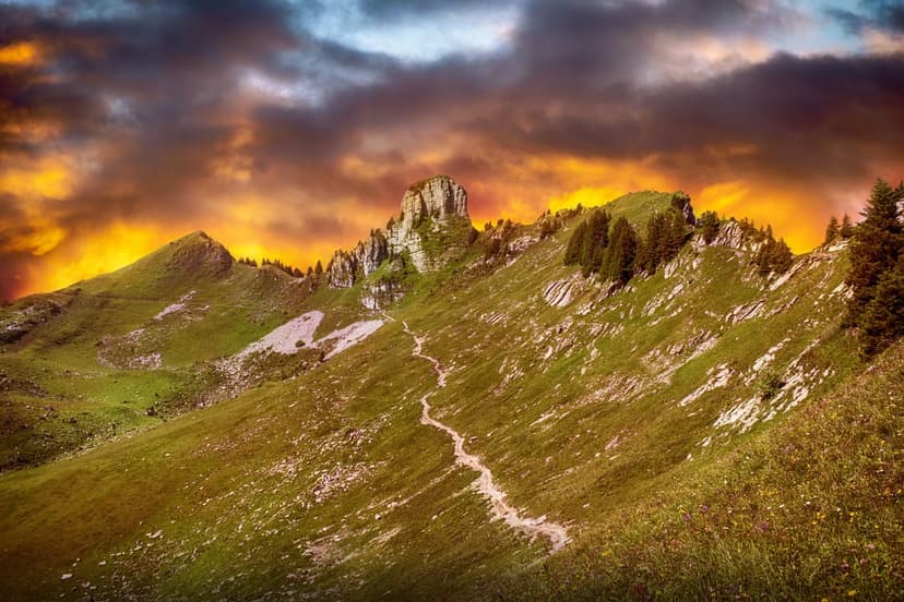 Jagged mountain peak under a purple and orange sky with a narrow hiking path through green grassy slopes.