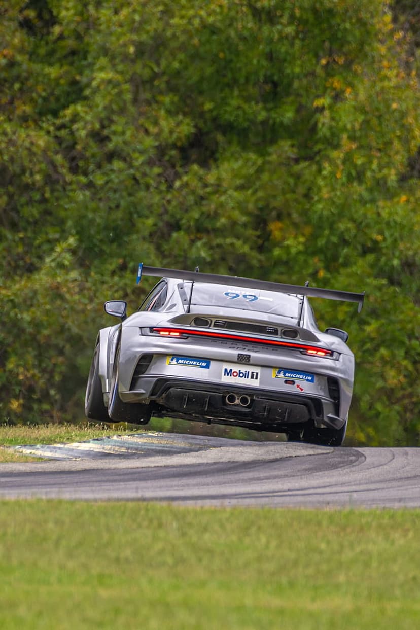 Silver Porsche GT3 racing car with number 99 navigates a track turn surrounded by lush green trees and grass.