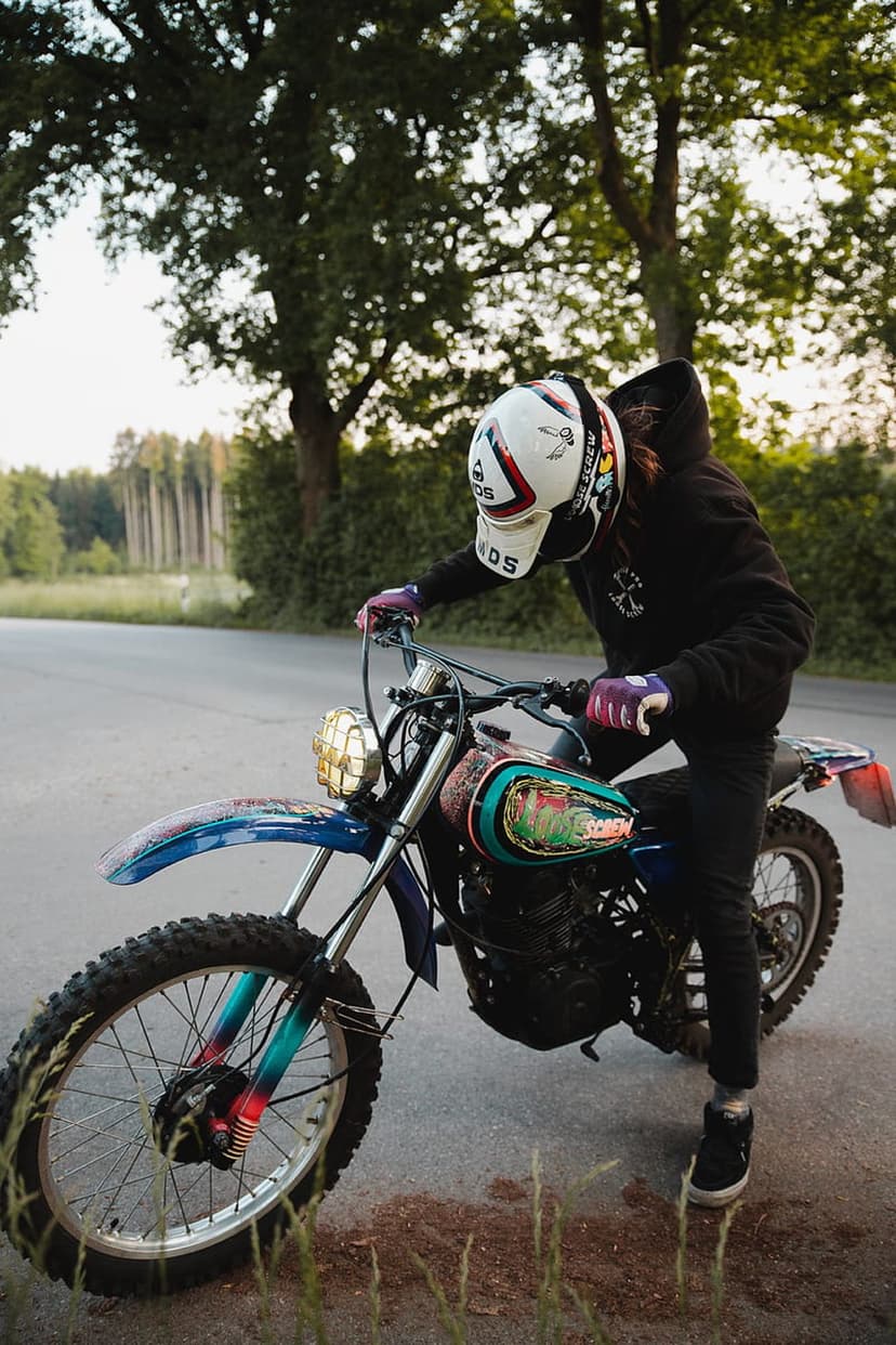 Motocross rider in a white helmet and black hoodie sits on a dirt bike with a colorful mural gas tank.