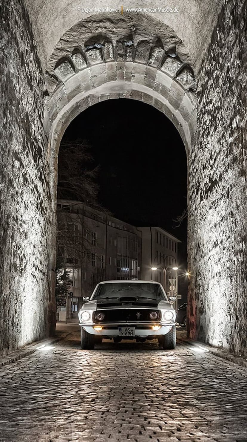 White Ford Mustang drives through a dark stone archway onto a wet cobblestone street at night under streetlights.