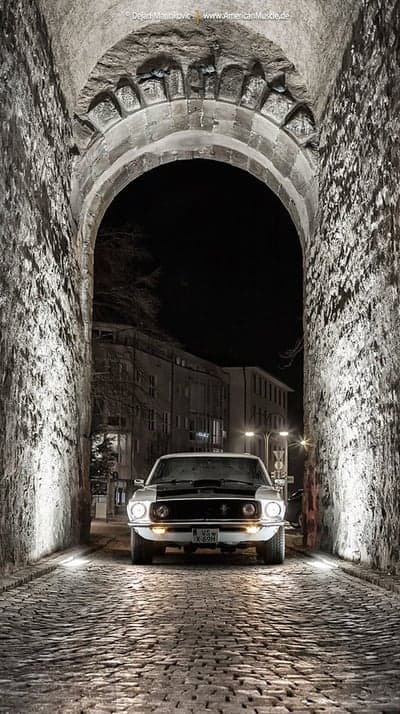 Classic White Mustang Drives Through Ancient Stone Archway at Night