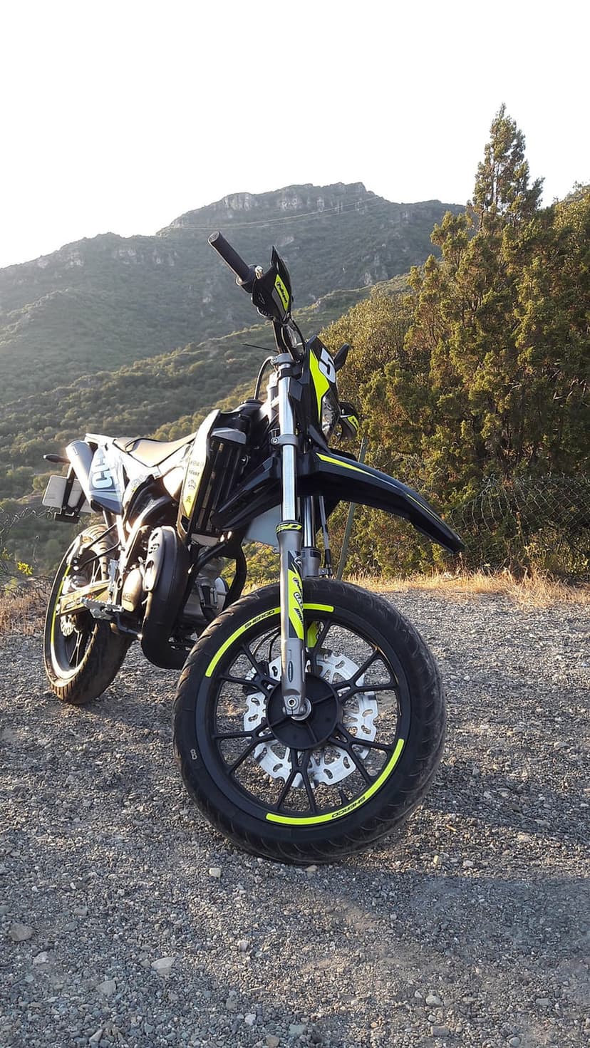 Supermoto motorcycle with yellow rims parked on a gravel mountain path overlooking pine trees and rocky peaks.