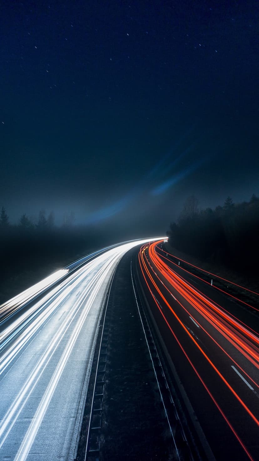 Luminous red and white light trails stretch along a forested highway under a dark starry night sky.