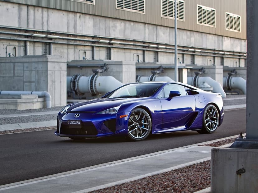 Deep blue Lexus LFA supercar parked on a road with industrial concrete buildings and large pipes in the background.