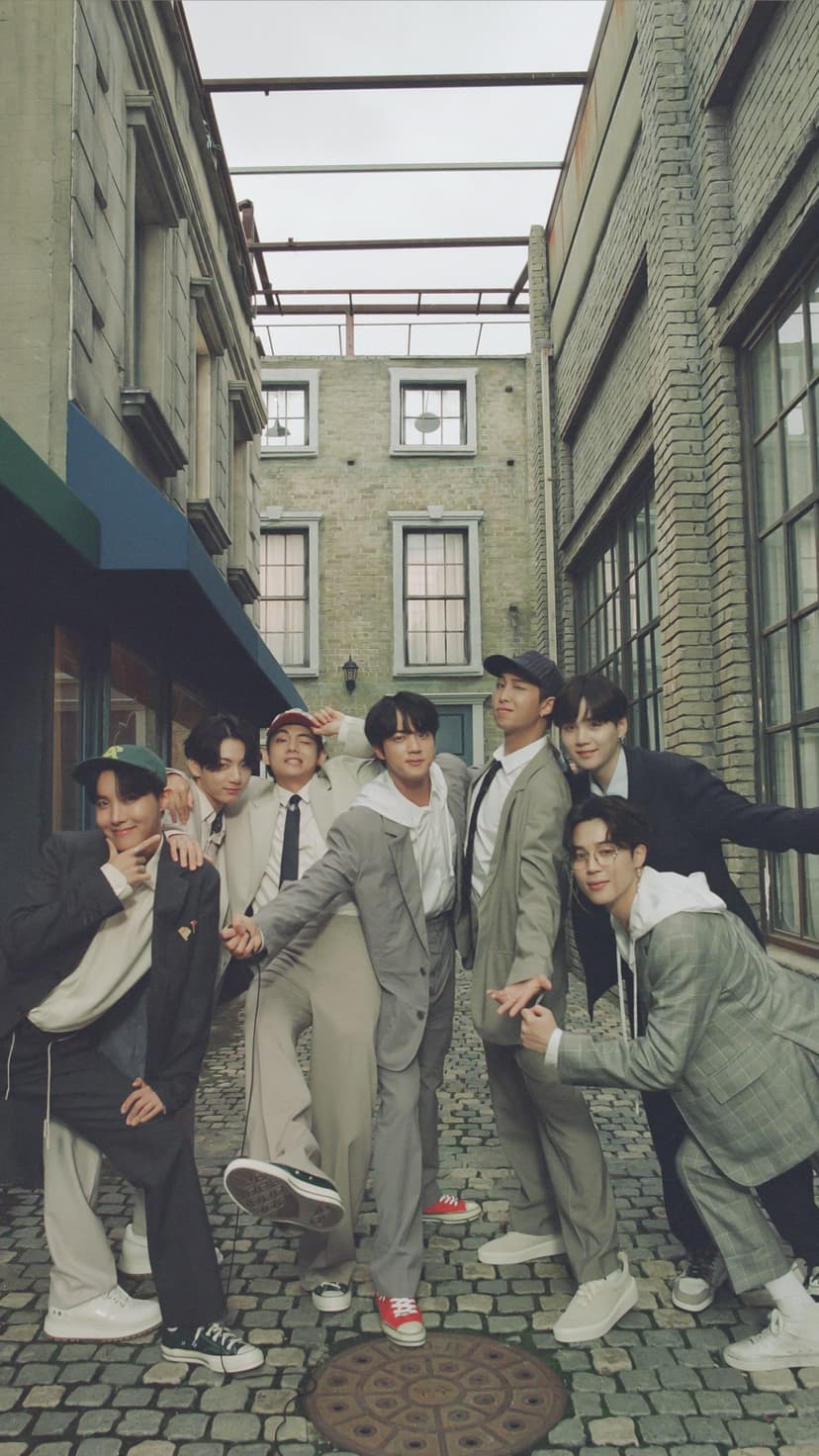 BTS members pose in streetwear within a brick alleyway wearing stylish jackets, caps, and leather sneakers.