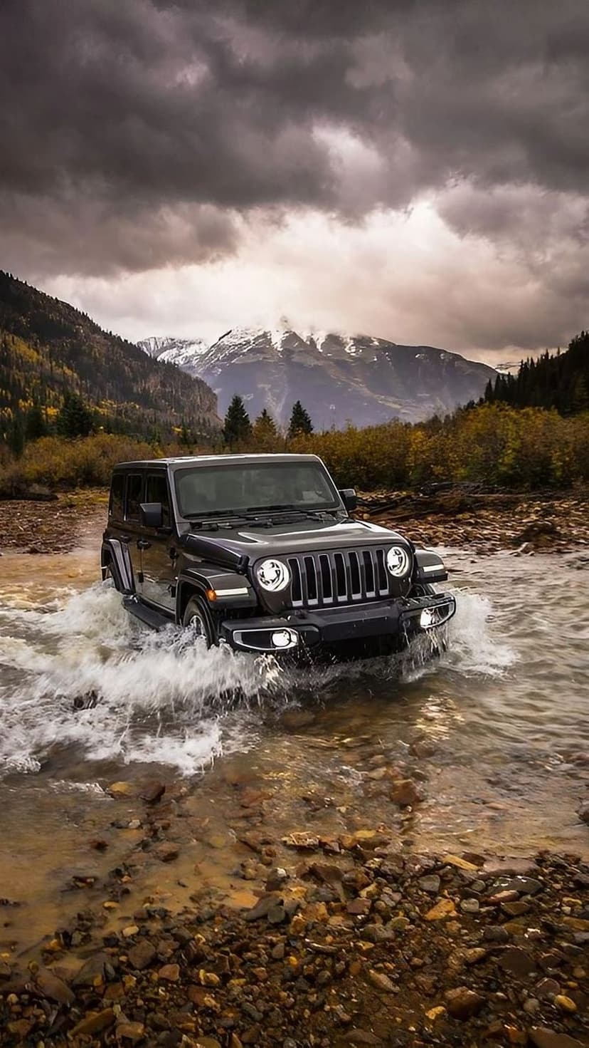 Jeep Wrangler splashing through a rocky mountain river under heavy storm clouds and distant snowy peaks.