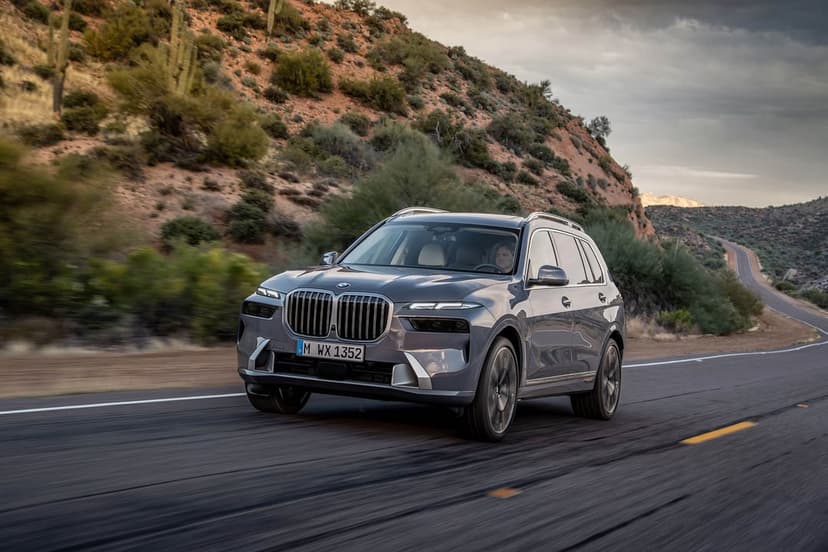 Grey BMW X7 SUV driving on a winding desert road past saguaro cacti and rocky hills under a cloudy sky.