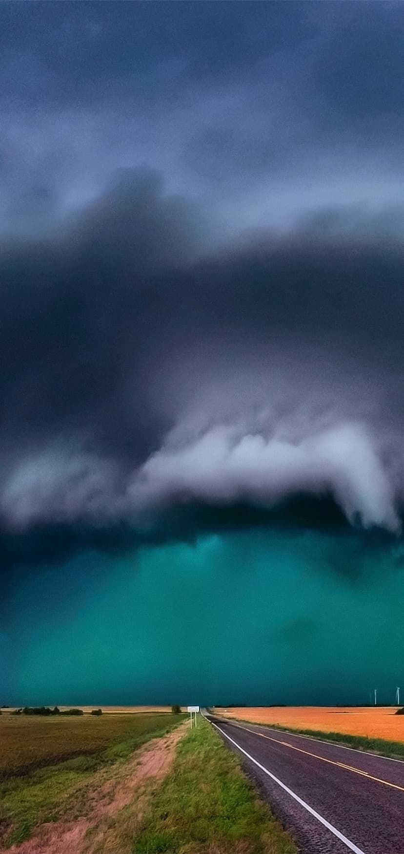Dark storm clouds hover over a straight asphalt road and a teal horizon in a dramatic rural landscape scene.