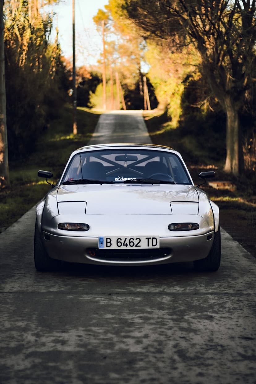 Front view of a silver widebody Mazda Miata NA with a roll cage on a narrow path under warm sunlight.