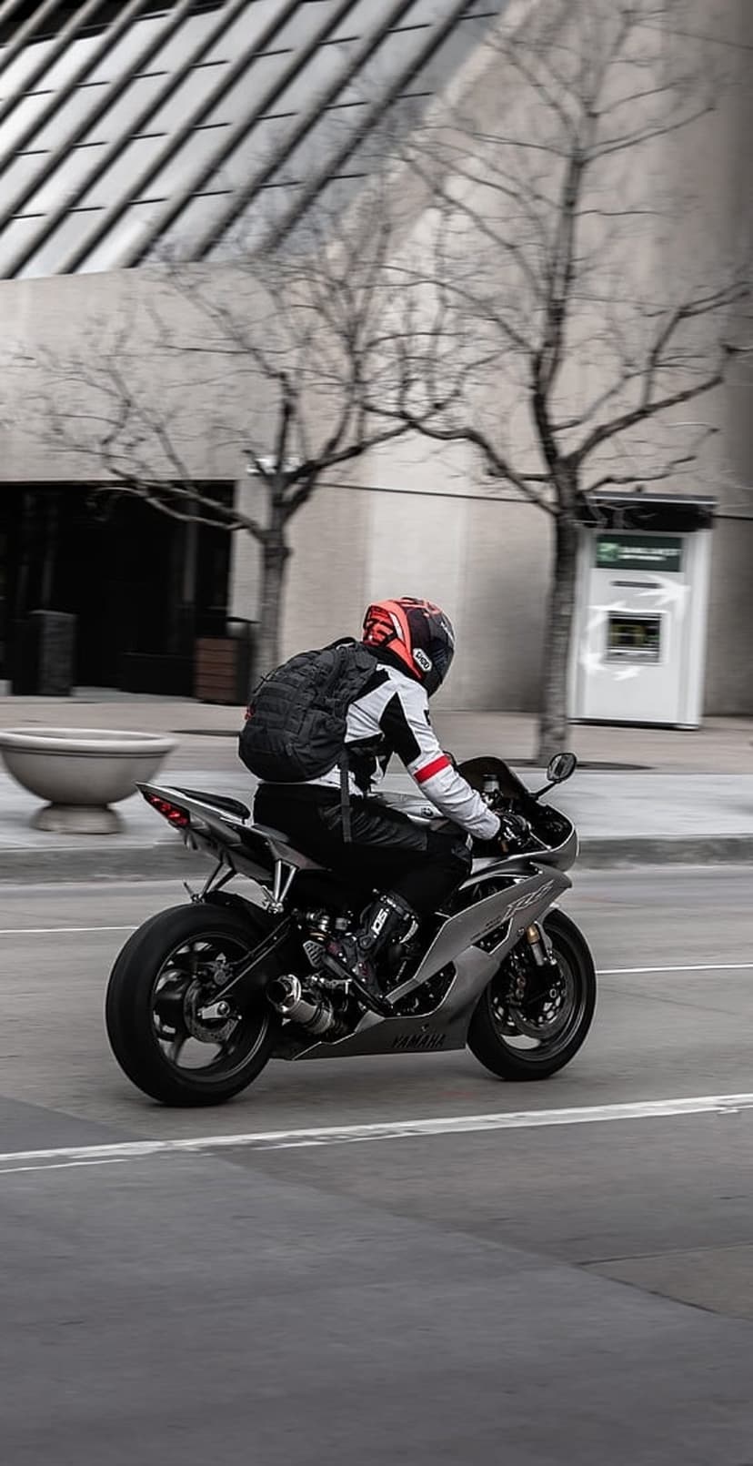 Motorcyclist in a silver helmet and backpack riding a sleek sportbike through a blurred urban street scene.
