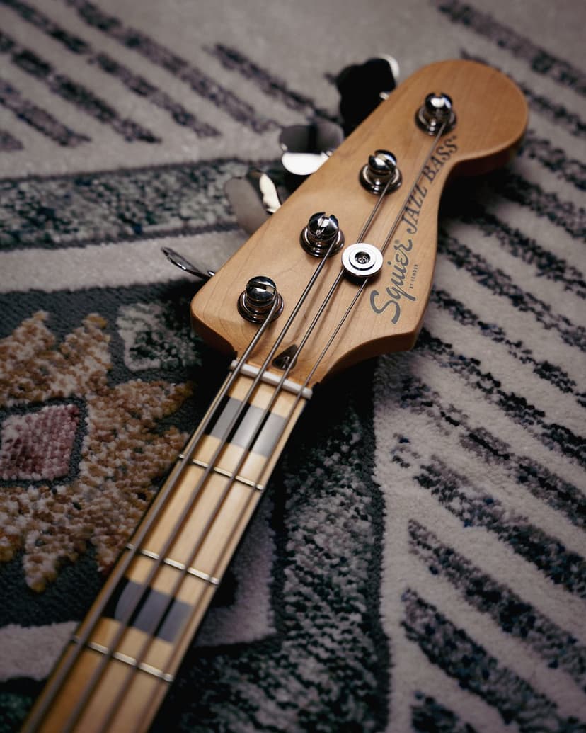 Squier Jazz Bass headstock with chrome tuning pegs and strings resting on a patterned rug in a close-up view.