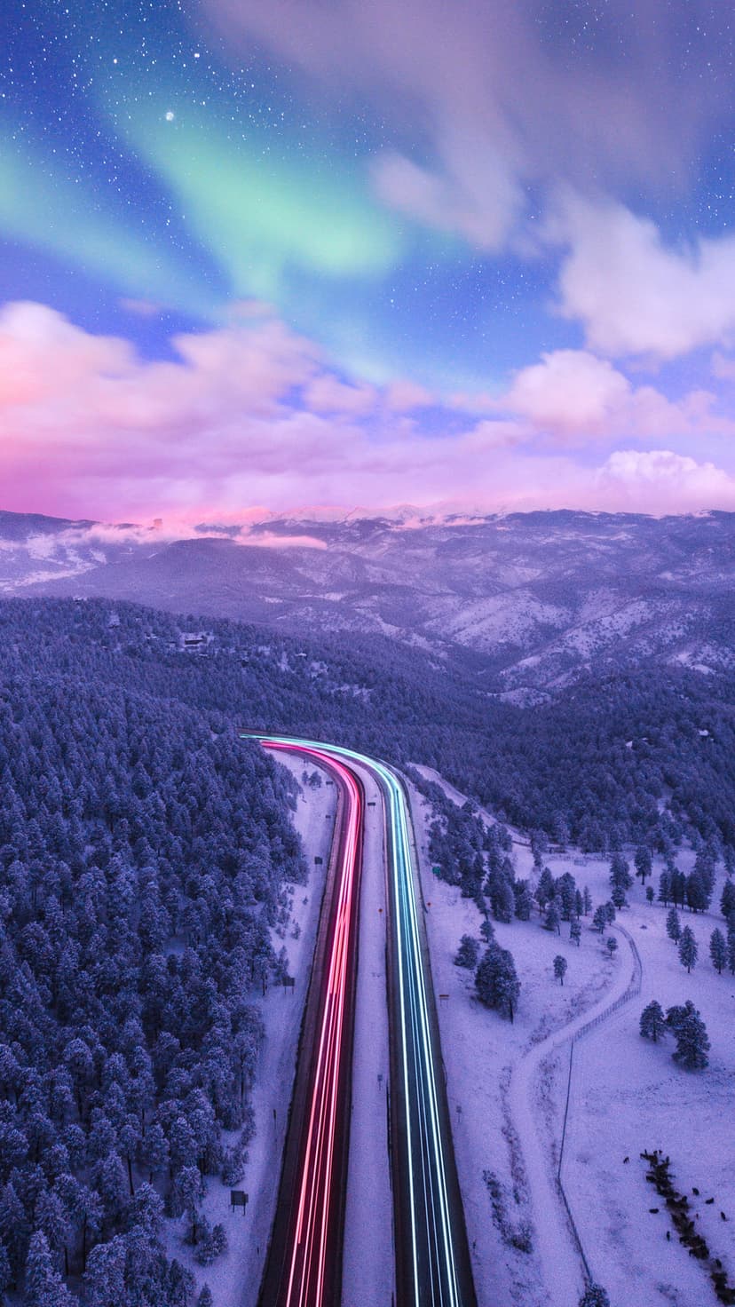 Winding snowy road with red and blue light trails through a pine forest under a pink and green aurora sky.