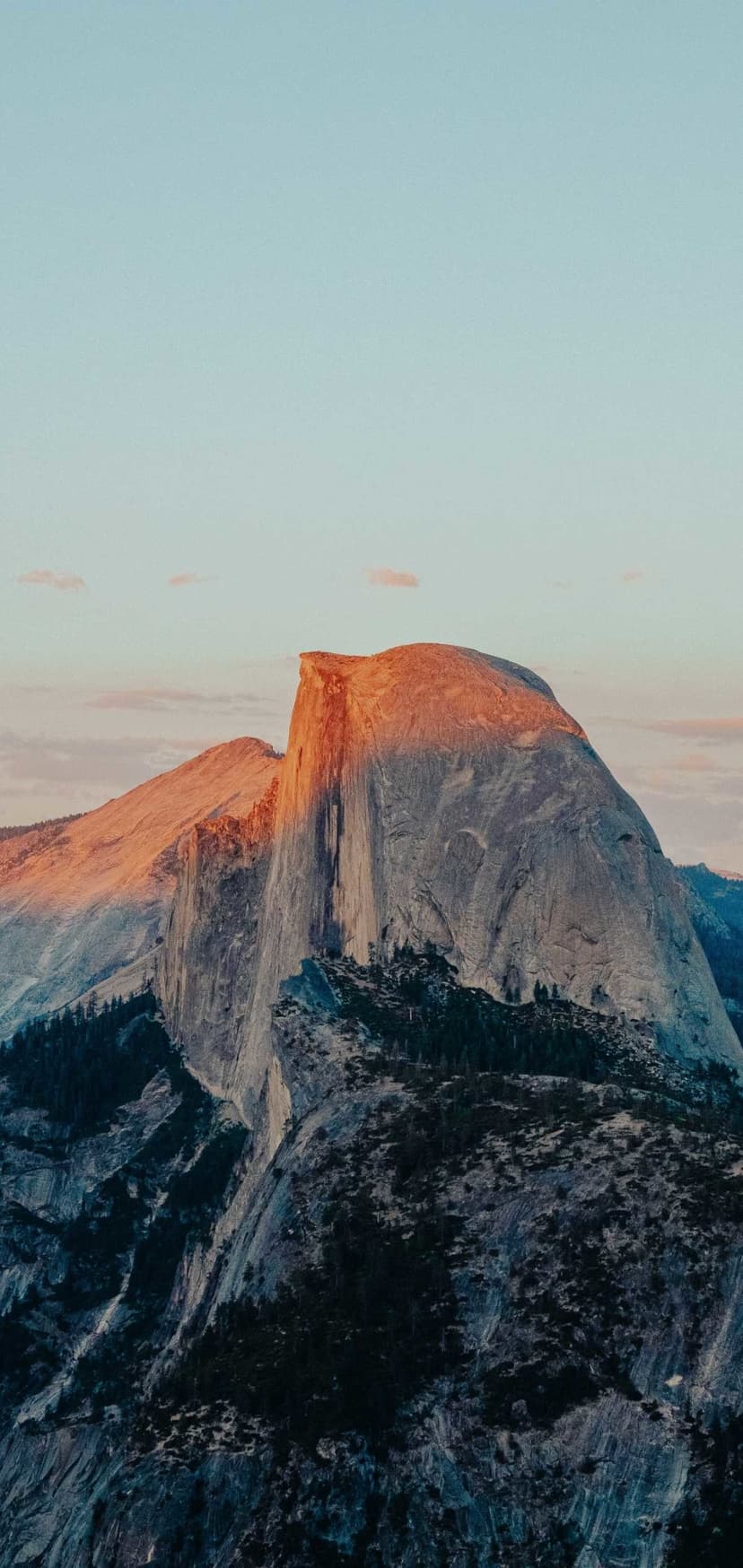 Half Dome's granite face glows orange at dusk above forested mountains under a soft pink and purple pastel sky.