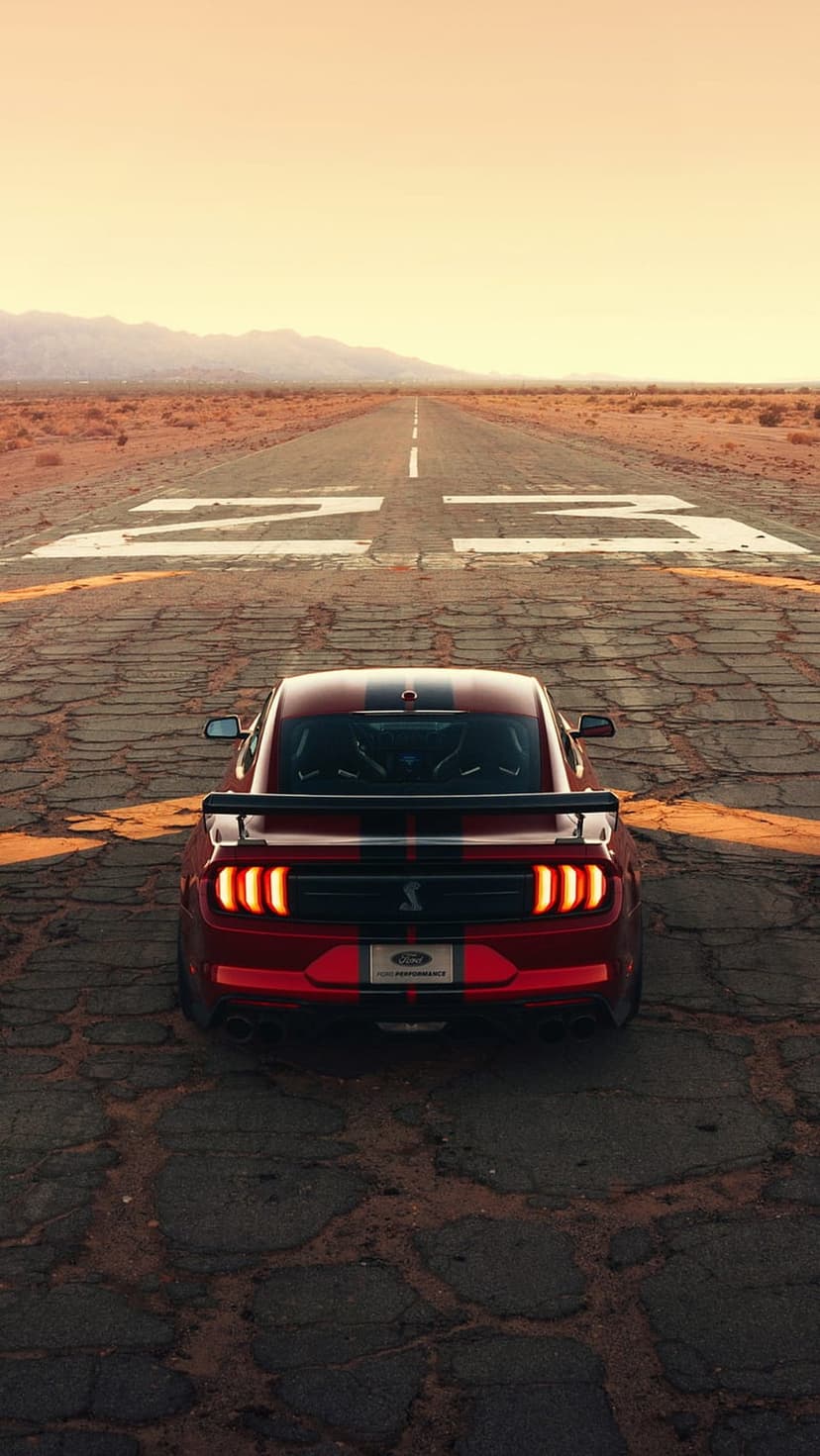 Red Shelby GT500 muscle car parked on a cracked desert runway under an orange sunset with glowing brake lights.