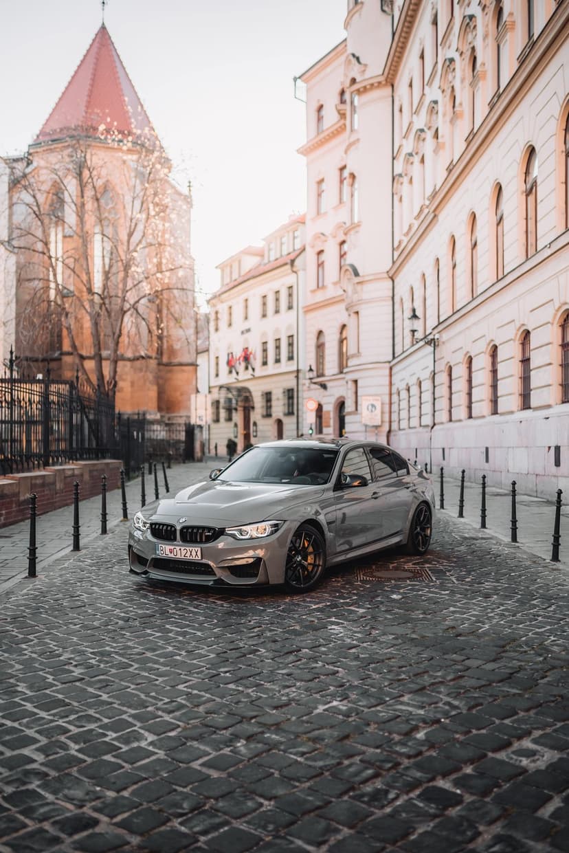 Gray BMW M4 parked on cobblestones with a historic church and old buildings under a warm sunset sky background.
