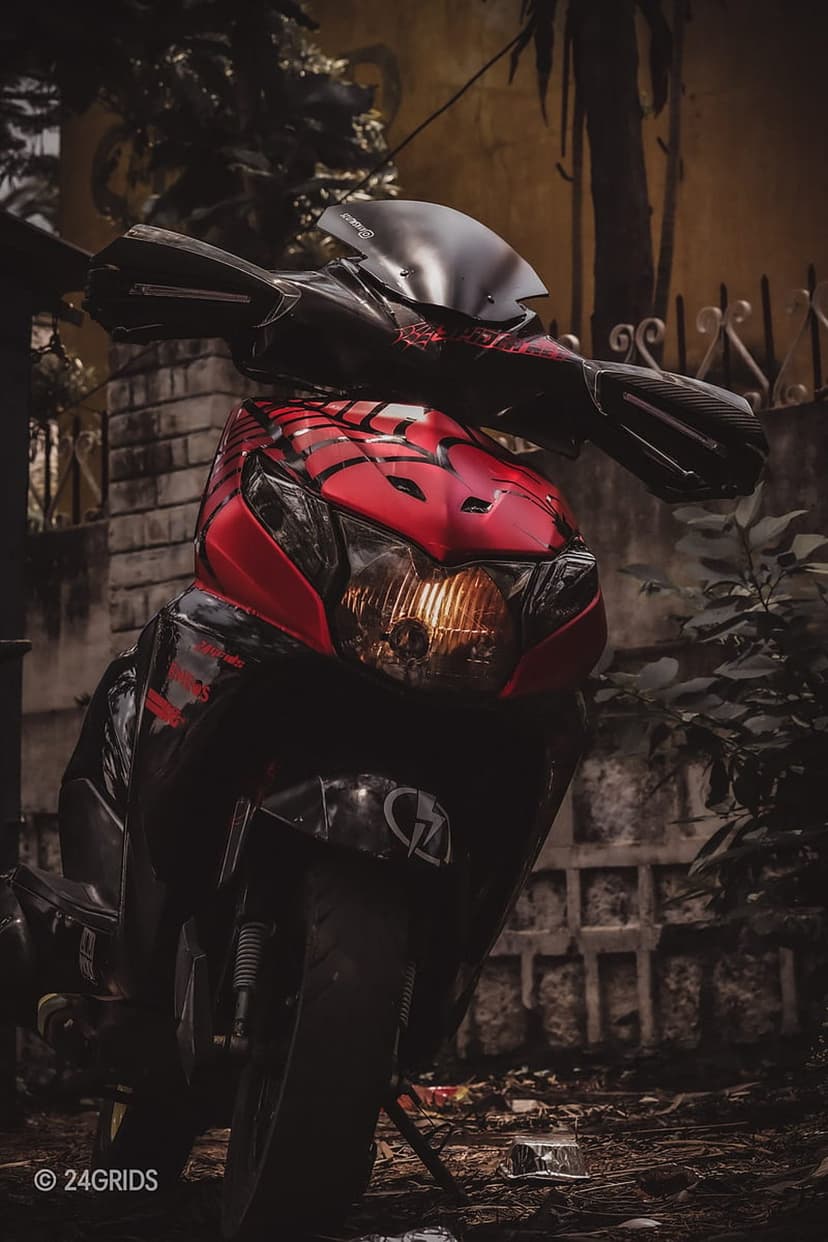 Red motorcycle with Spider-Man decals and glowing headlights parked against a dark brick and leaf background.