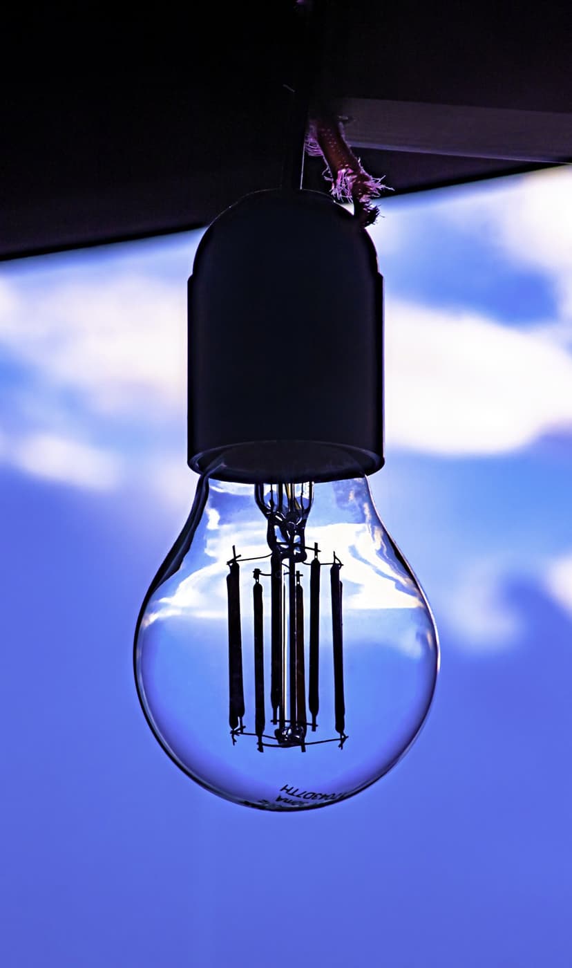 Vintage glass light bulb with visible filament hanging against a vibrant blue sky filled with white fluffy clouds.