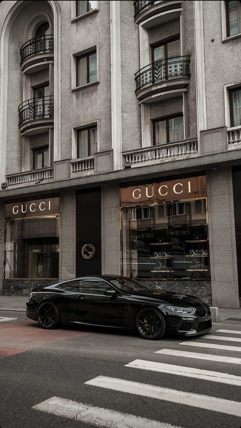 Black luxury sports car parked outside a Gucci boutique with classical architecture and gold shop lighting.