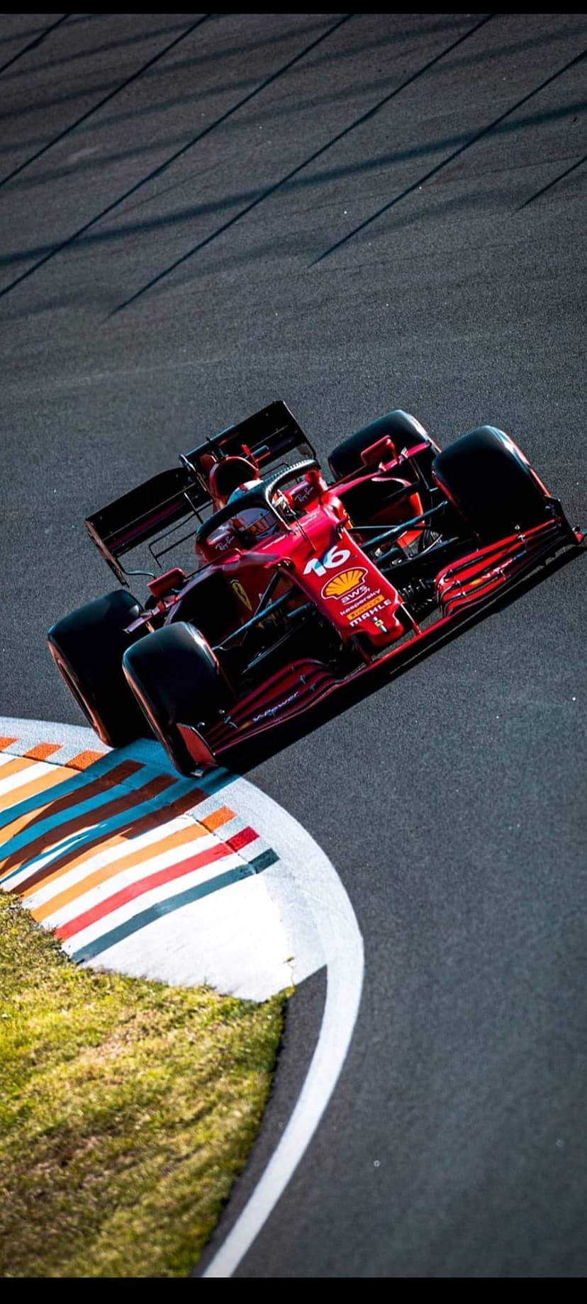 Ferrari F1 car with number 16 racing through a track corner with colorful curbing and green grass embankments.