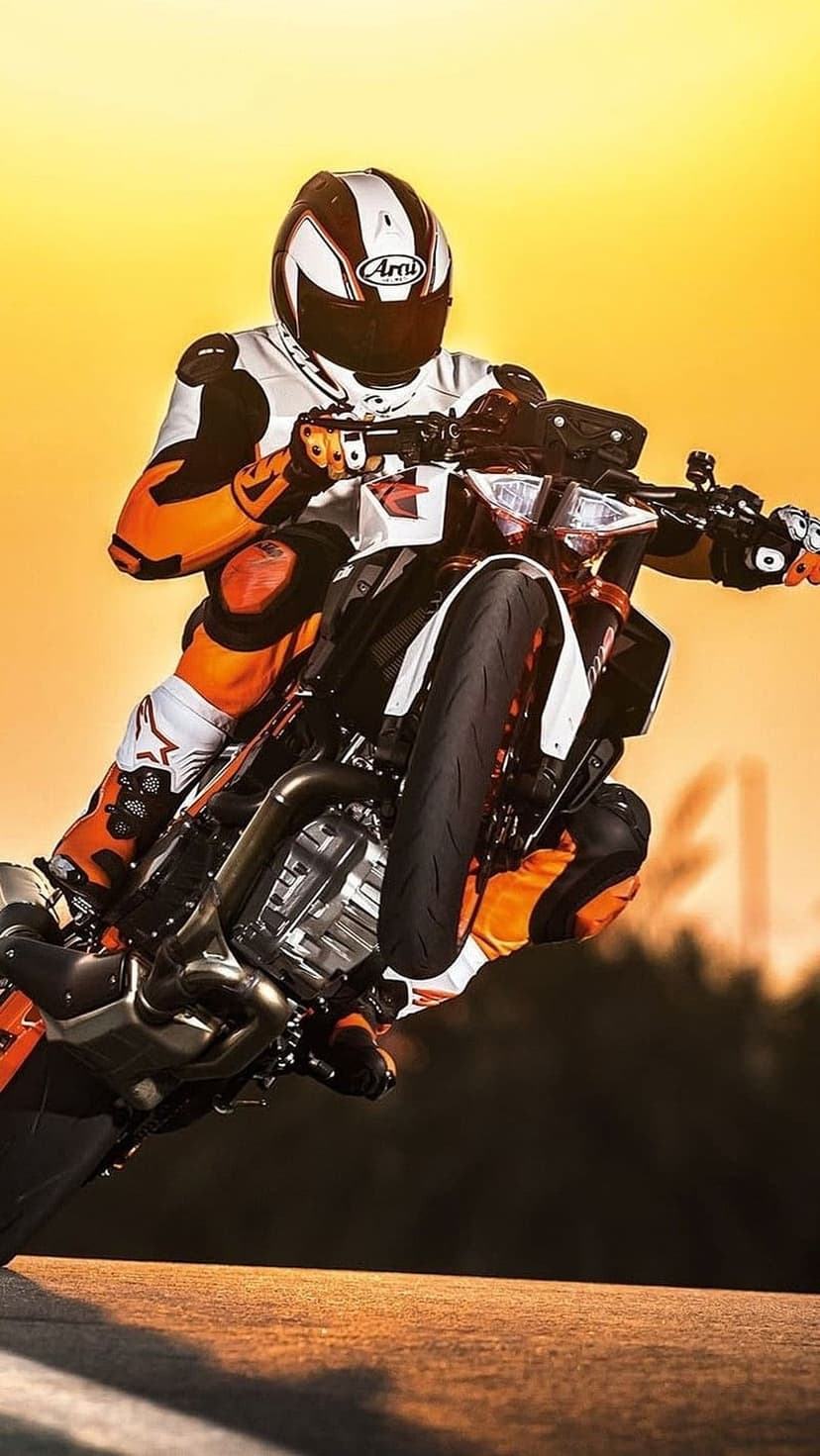 Motorcyclist in orange and white racing gear performs a wheelie on a sportbike during a vibrant orange sunset.