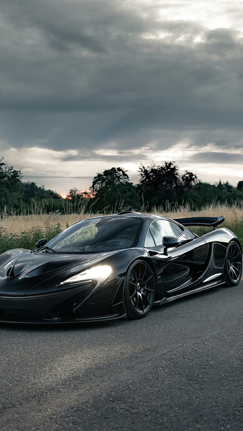 Black McLaren P1 supercar with glowing headlights parked on a rural road under a dark twilight sky at evening.