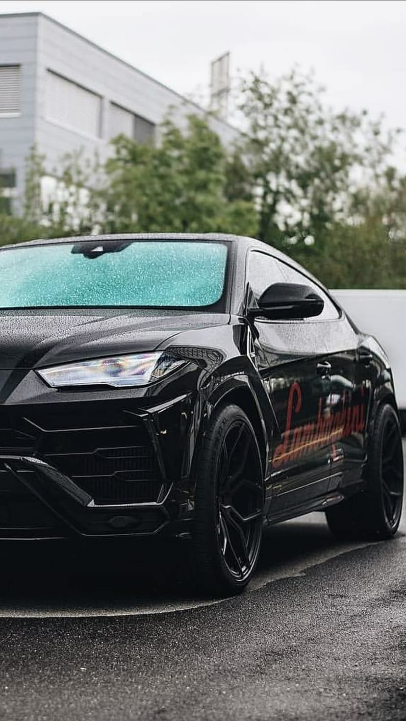Black Lamborghini Urus SUV parked on a wet asphalt street with raindrops on the glass and urban reflections.