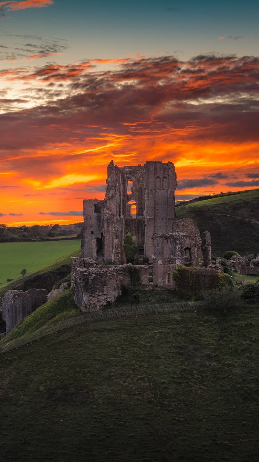 Corfe Castle stone ruins sit atop a green hill under a dramatic sky of orange and red clouds at sunset.
