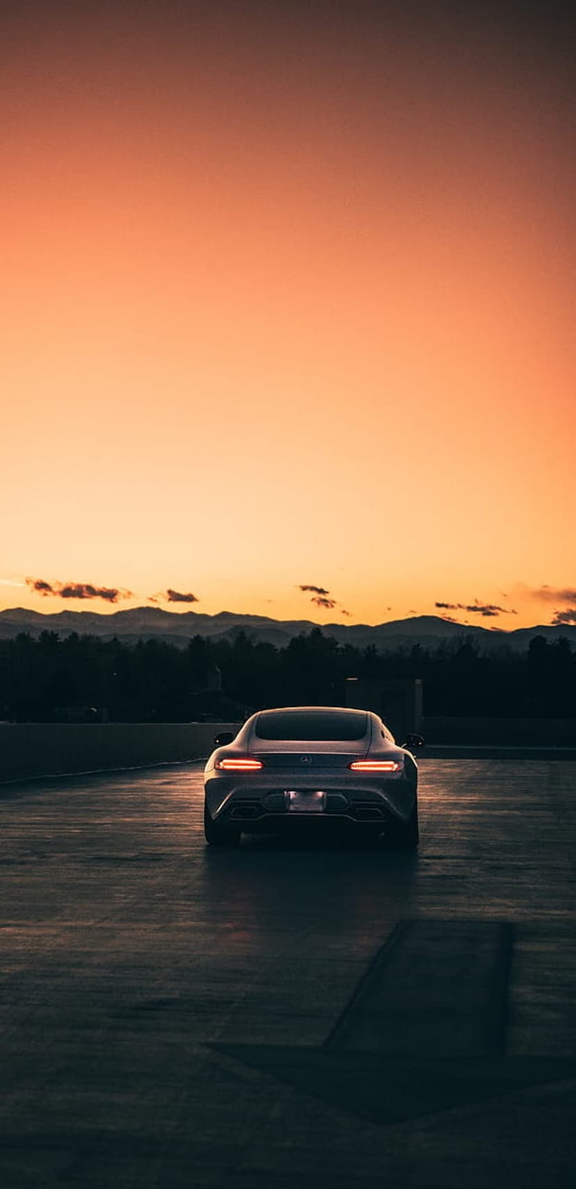 Luxury sports car with red taillights parked on asphalt during a mountain sunset with an orange and purple sky.
