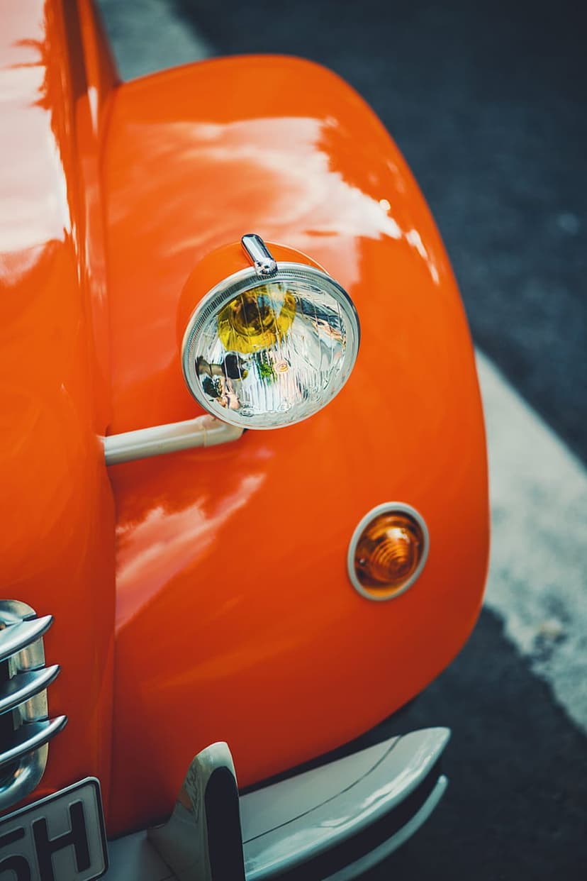 Vintage orange car fender and circular chrome headlight with a yellow bulb reflect clouds on a glossy surface.