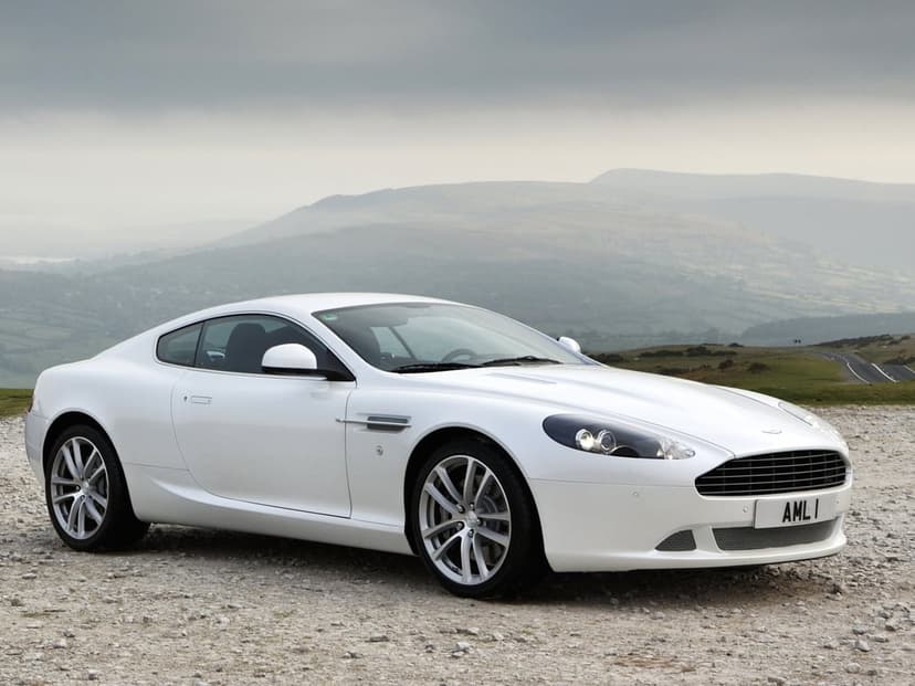 White luxury sports car parked on a gravel overlook with misty hills and an overcast sky in the background.